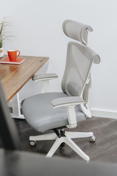 An ergonomic office chair with a high mesh backrest and adjustable headrest is positioned next to a wooden desk. The desk has a plant and a red mug on it, as well as a closed red notebook.