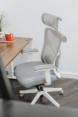 An ergonomic office chair with a high mesh backrest and adjustable headrest is positioned next to a wooden desk. The desk has a plant and a red mug on it, as well as a closed red notebook.