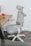 An ergonomic office chair with a high mesh backrest and adjustable headrest is positioned next to a wooden desk. The desk has a plant and a red mug on it, as well as a closed red notebook.