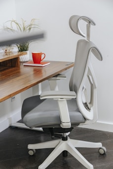 A modern ergonomic office chair with a gray cushioning sits beside a wooden desk. On the desk, there is a red coffee mug and a red notebook. A small potted plant adds a touch of nature to the workspace.
