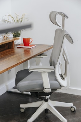 A modern ergonomic office chair with a gray cushioning sits beside a wooden desk. On the desk, there is a red coffee mug and a red notebook. A small potted plant adds a touch of nature to the workspace.