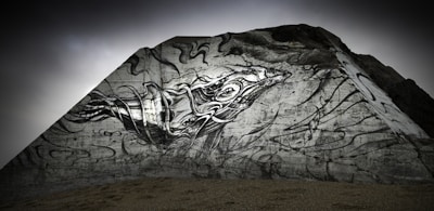A dramatic mural painted on a large triangular rock structure, featuring an abstract, stylized depiction of a sea creature. The image includes dynamic lines and swirling patterns, contributing to its sense of movement and flow. The background is overcast, adding a mysterious atmosphere.