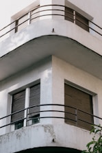 A modern architectural building featuring a corner with curved balconies. The structure is predominantly white, with two levels of balconies encased by metal railings. The windows have closed shutters, adding a sense of privacy. The building's exterior shows signs of weathering.