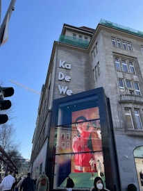 An urban street scene featuring a large building adorned with the letters 'Ka De We'. A prominent digital advertisement display is visible, showcasing a fashion model in vivid colors. People are walking by, some wearing winter clothing and masks. The sky is clear with a construction crane in the background.