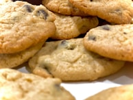 A close-up view of freshly baked chocolate chip cookies with a golden-brown color, displaying a textured surface and visible chocolate chips. The image captures the cookies at an angle, emphasizing their round shape and slightly crumbled edges.