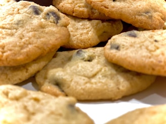 A close-up view of freshly baked chocolate chip cookies with a golden-brown color, displaying a textured surface and visible chocolate chips. The image captures the cookies at an angle, emphasizing their round shape and slightly crumbled edges.
