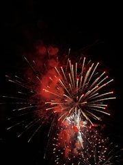 A vibrant display of fireworks with bright red and white bursts, set against the dark night sky. The fireworks are exploding in various shapes and sizes, creating a dazzling spectacle.
