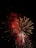 A vibrant display of fireworks with bright red and white bursts, set against the dark night sky. The fireworks are exploding in various shapes and sizes, creating a dazzling spectacle.