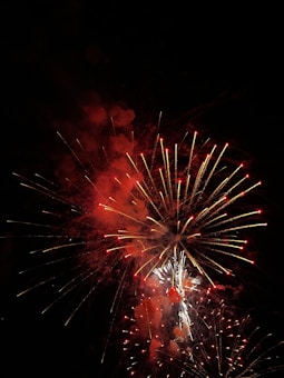 A vibrant display of fireworks with bright red and white bursts, set against the dark night sky. The fireworks are exploding in various shapes and sizes, creating a dazzling spectacle.