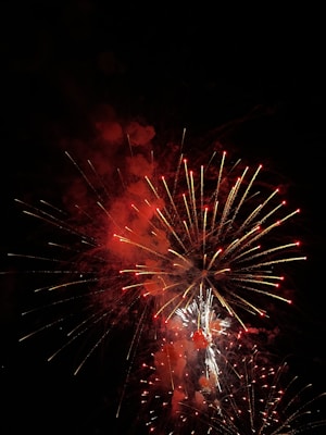A vibrant display of fireworks with bright red and white bursts, set against the dark night sky. The fireworks are exploding in various shapes and sizes, creating a dazzling spectacle.