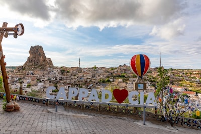 A picturesque view features the rocky terrain and unique rock formations of Cappadocia, with a sprawling town in the foreground. A large decorative sign with the word 'CAPPADOCIA' and a heart symbol can be seen, accompanied by a colorful hot air balloon structure. The sky is partly cloudy, adding depth to the scene, while decorative elements like charms and colorful cloths enhance the foreground.