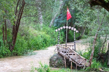 A small wooden bridge extends over a narrow, murky stream surrounded by dense green foliage. Above the bridge, there is an arched wooden structure adorned with hanging signs spelling 'BELISIRMA' and a red flag with a white crescent and star. The scene is peaceful, nestled within a natural setting.