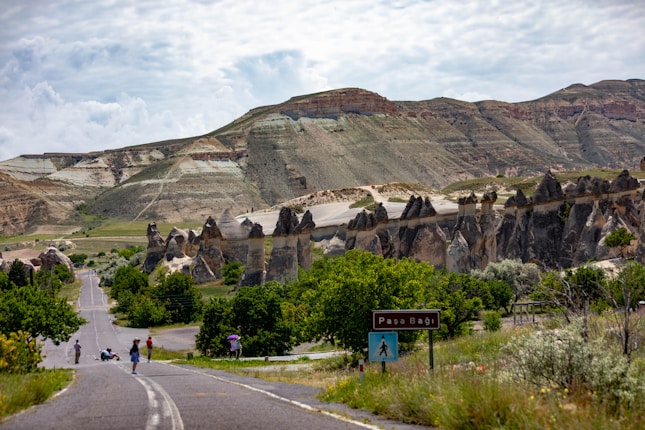 A picturesque landscape features unique rock formations against a backdrop of rolling hills and a partly cloudy sky. A road winds through the scene, bordered by lush greenery. Several people are visible, some walking and others standing, near a brown signpost that includes the text 'Paşa Bağı'.