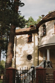 A classical-style building with arched windows and a round, ornamented window. The structure features ornate white columns and a curved staircase leading to the entrance. Tall trees surround the area, casting shadows across the cream-colored walls of the building.