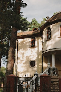 A classical-style building with arched windows and a round, ornamented window. The structure features ornate white columns and a curved staircase leading to the entrance. Tall trees surround the area, casting shadows across the cream-colored walls of the building.