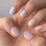 A close-up of a hand showing manicured nails painted in soft pastel shades, including light pink, pale yellow, and gentle green. The nails have a glossy finish, and the skin tone is warm and natural.