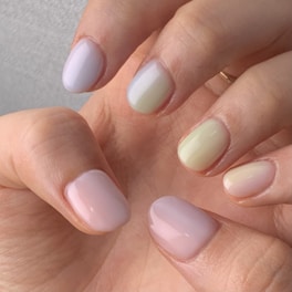 A close-up of a hand showing manicured nails painted in soft pastel shades, including light pink, pale yellow, and gentle green. The nails have a glossy finish, and the skin tone is warm and natural.