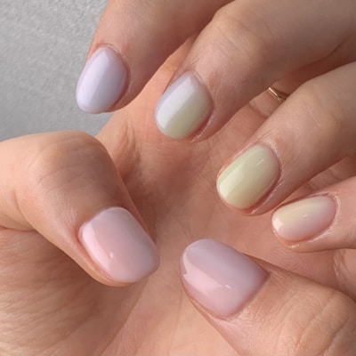 A close-up of a hand showing manicured nails painted in soft pastel shades, including light pink, pale yellow, and gentle green. The nails have a glossy finish, and the skin tone is warm and natural.