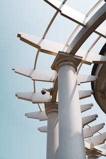 A modern architectural structure with tall round columns supporting a circular roof structure with open slats, creating a geometric pattern against the clear sky. A surveillance camera is mounted near the top of one column, adding an element of technology.
