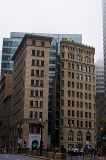 Two historical buildings with intricate architecture stand prominently in front of a modern glass skyscraper. The older structures feature detailed windows and ornate design elements. Pedestrians walk along the street in front of the buildings. A sign for Brookline Bank is visible at the base of one of the structures.