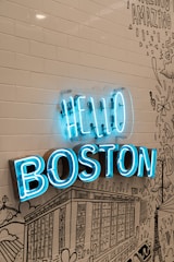 A vibrant blue neon sign reads 'HELLO BOSTON' mounted on a wall with white subway tiles and black line art illustrations depicting cityscapes and various designs.