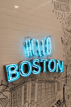 A vibrant blue neon sign reads 'HELLO BOSTON' mounted on a wall with white subway tiles and black line art illustrations depicting cityscapes and various designs.