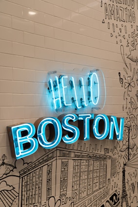 A vibrant blue neon sign reads 'HELLO BOSTON' mounted on a wall with white subway tiles and black line art illustrations depicting cityscapes and various designs.