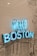 A vibrant blue neon sign reads 'HELLO BOSTON' mounted on a wall with white subway tiles and black line art illustrations depicting cityscapes and various designs.