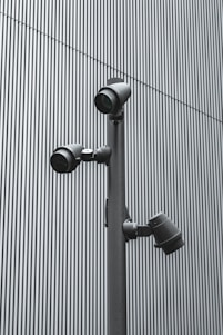 A trio of modern security cameras attached to a vertical metal pole, set against a backdrop of gray vertical lines, evoking an industrial and surveillance-themed setting.