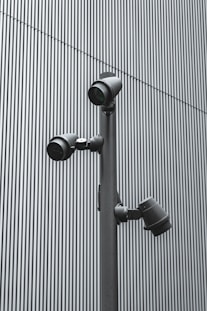 A trio of modern security cameras attached to a vertical metal pole, set against a backdrop of gray vertical lines, evoking an industrial and surveillance-themed setting.