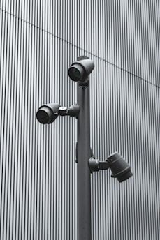 A trio of modern security cameras attached to a vertical metal pole, set against a backdrop of gray vertical lines, evoking an industrial and surveillance-themed setting.