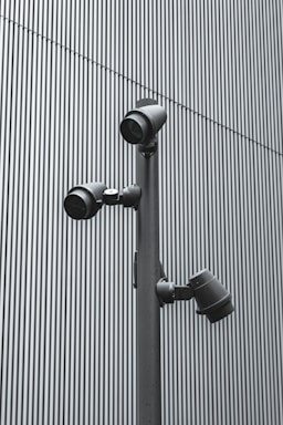 A trio of modern security cameras attached to a vertical metal pole, set against a backdrop of gray vertical lines, evoking an industrial and surveillance-themed setting.
