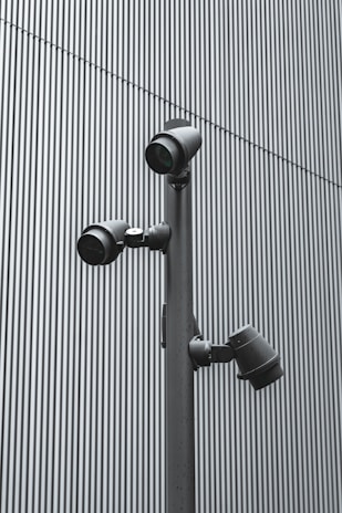 A trio of modern security cameras attached to a vertical metal pole, set against a backdrop of gray vertical lines, evoking an industrial and surveillance-themed setting.