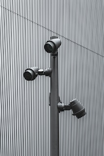 A trio of modern security cameras attached to a vertical metal pole, set against a backdrop of gray vertical lines, evoking an industrial and surveillance-themed setting.