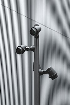A trio of modern security cameras attached to a vertical metal pole, set against a backdrop of gray vertical lines, evoking an industrial and surveillance-themed setting.