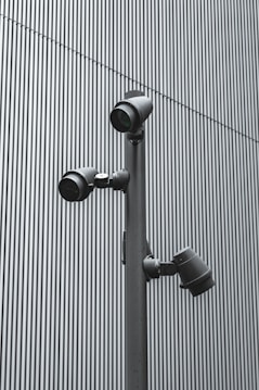 A trio of modern security cameras attached to a vertical metal pole, set against a backdrop of gray vertical lines, evoking an industrial and surveillance-themed setting.