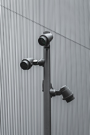 A trio of modern security cameras attached to a vertical metal pole, set against a backdrop of gray vertical lines, evoking an industrial and surveillance-themed setting.