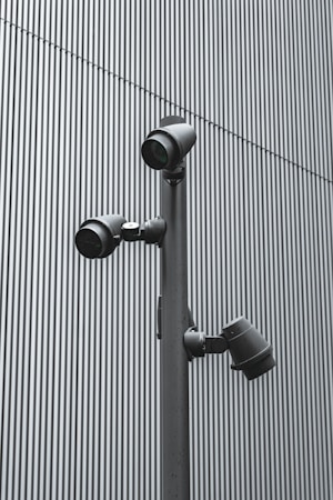 A trio of modern security cameras attached to a vertical metal pole, set against a backdrop of gray vertical lines, evoking an industrial and surveillance-themed setting.