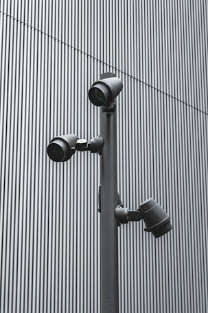 A trio of modern security cameras attached to a vertical metal pole, set against a backdrop of gray vertical lines, evoking an industrial and surveillance-themed setting.