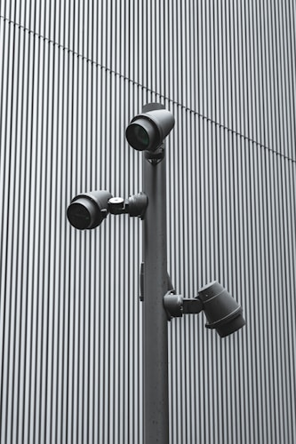 A trio of modern security cameras attached to a vertical metal pole, set against a backdrop of gray vertical lines, evoking an industrial and surveillance-themed setting.