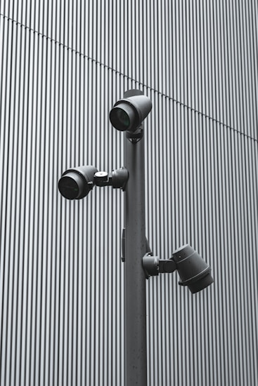A trio of modern security cameras attached to a vertical metal pole, set against a backdrop of gray vertical lines, evoking an industrial and surveillance-themed setting.