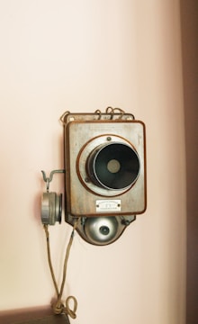 A vintage wall-mounted telephone with a wooden casing and a large central speaker. It features a metal earpiece connected by a cord and intricate metal fittings. The background is a plain light-colored wall.
