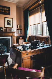 A vintage office setting featuring a wooden desk with an antique typewriter, surrounded by classic furniture and decor. The room has large windows with partially drawn blinds, soft light filtering through. A portrait hangs above a fireplace, and books are neatly arranged on a mantelpiece. Dark green curtains frame the windows, complementing the rich wooden textures of the room.