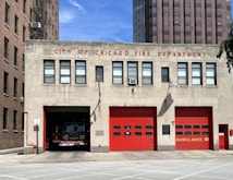A fire station with a sign reading 'City of Chicago Fire Department' at the top. It features a red fire truck partially visible in one of the open garage doors. There are three red garage doors in total, one labeled 'Ambulance 41'. The building is flanked by tall urban structures, and the scene is lit by bright daylight with clear skies.