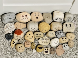 A collection of smooth stones arranged on a textured surface, each featuring a painted face displaying a variety of emotions including happiness, sadness, surprise, and anger. The stones are of different shapes, sizes, and colors, creating a playful and whimsical assortment.