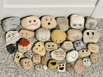 A collection of smooth stones arranged on a textured surface, each featuring a painted face displaying a variety of emotions including happiness, sadness, surprise, and anger. The stones are of different shapes, sizes, and colors, creating a playful and whimsical assortment.