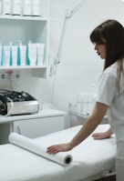 A woman is preparing a treatment table in what appears to be a spa or medical setting. She is adjusting a sheet or roll of paper on the table. Shelves behind her are stocked with various bottles, likely skincare or medical products. A large adjustable lamp is attached to the workspace.
