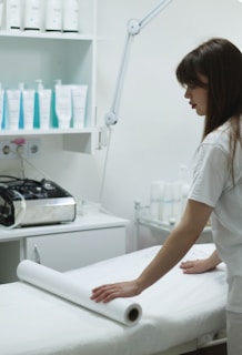 A woman is preparing a treatment table in what appears to be a spa or medical setting. She is adjusting a sheet or roll of paper on the table. Shelves behind her are stocked with various bottles, likely skincare or medical products. A large adjustable lamp is attached to the workspace.