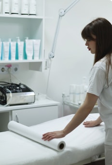 A woman is preparing a treatment table in what appears to be a spa or medical setting. She is adjusting a sheet or roll of paper on the table. Shelves behind her are stocked with various bottles, likely skincare or medical products. A large adjustable lamp is attached to the workspace.