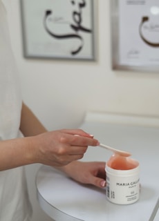 A person is using a spatula to scoop a pink gel-like substance from a container labeled 'Maria Galland Paris'. The person is wearing a white shirt, and the background includes abstract artwork and a white table.